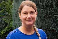 Person mit geflochtenem Zopf und blauem Oberteil. Im Hintergrund sind grüne Bäume zu sehen.